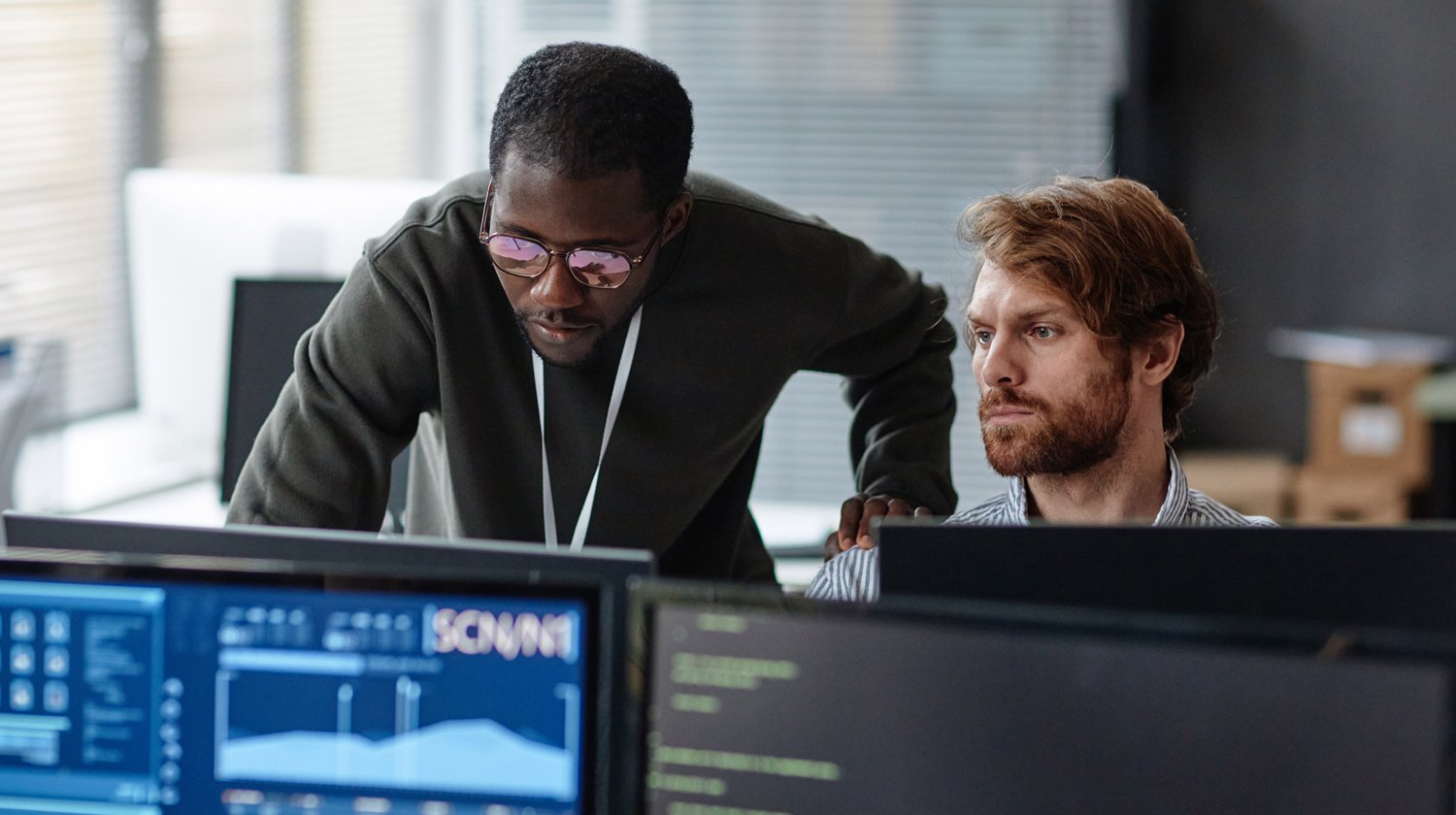 Two focused male colleagues analyze computer screens in an office. The man on the left leans over, wearing glasses, while the seated man listens intently.