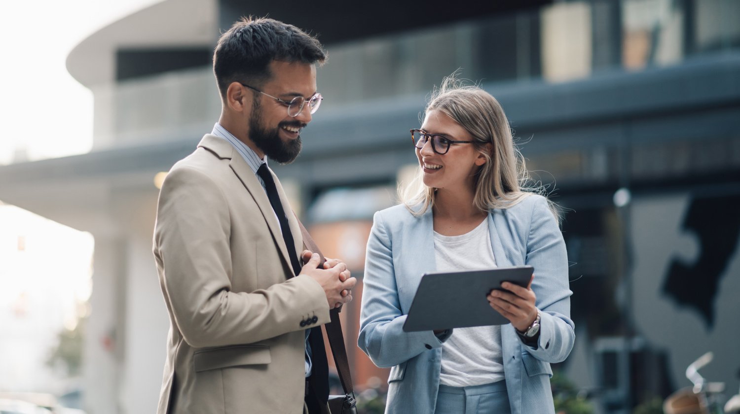 A man and woman in business attire happily discuss outside. The woman holds a tablet, both smiling, suggesting a professional, collaborative atmosphere.