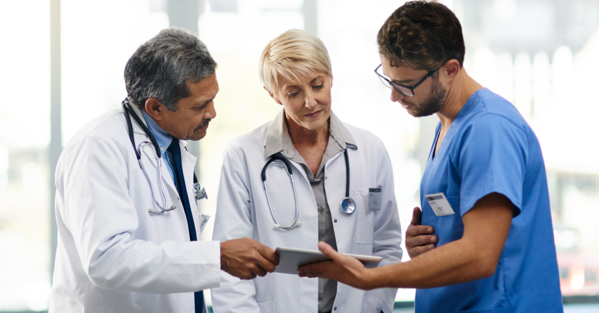 Three medical professionals, two in white coats and one in blue scrubs, discuss information displayed on a digital tablet in a hospital setting