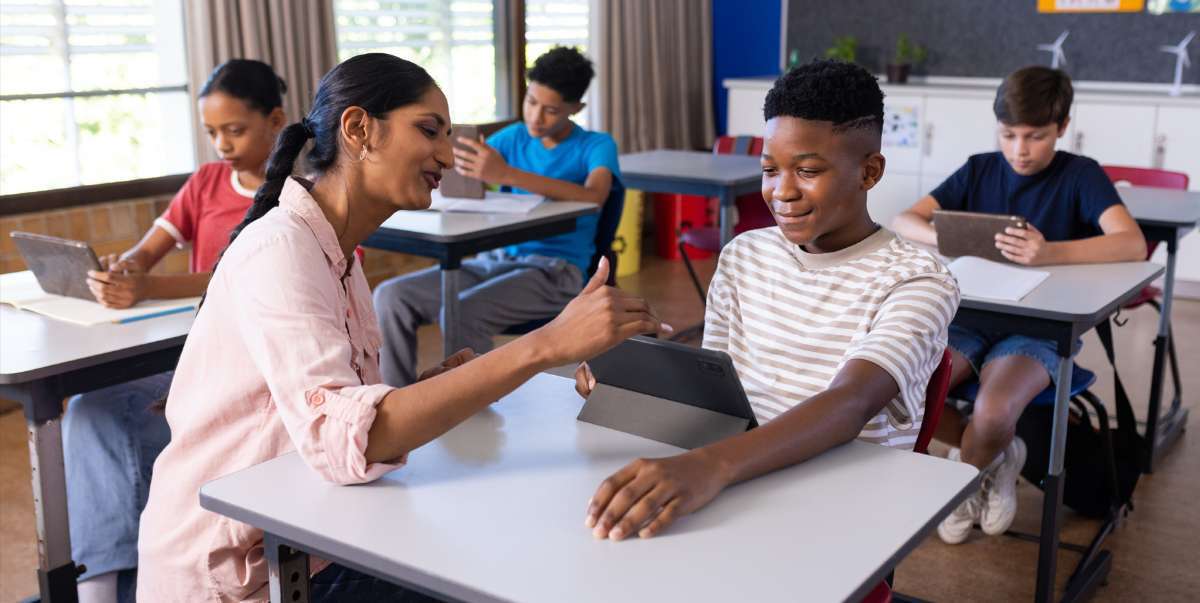 Teacher interacting with a smiling student who is using a tablet in a classroom, while other students in the background also use tablets at their desks.